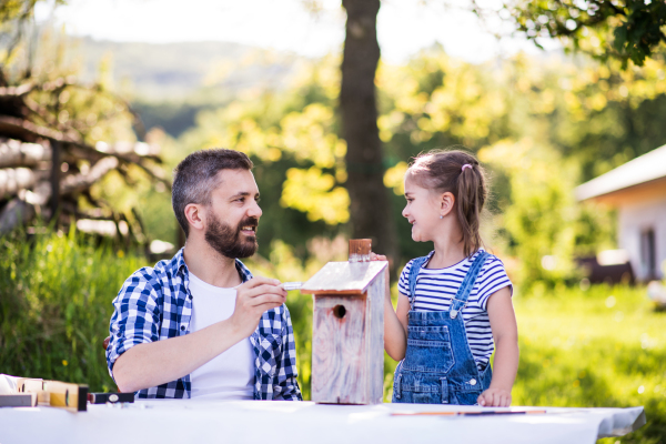 Mature father with a small daughter outside, painting. Wooden birdhouse or bird feeder making.