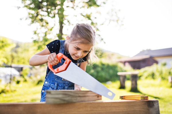 A happy small girl with a saw outside, making a wooden birdhouse or bird feeder.