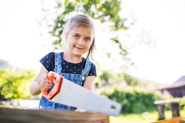 A happy small girl with a saw outside, making a wooden birdhouse or bird feeder.