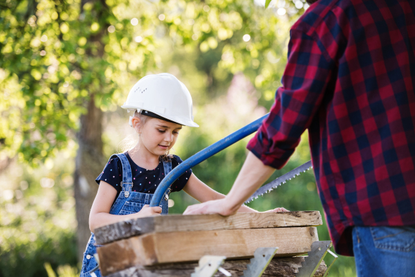 Unrecognizable father and a small daughter outside with a saw, cutting wood. Wooden birdhouse or bird feeder making.