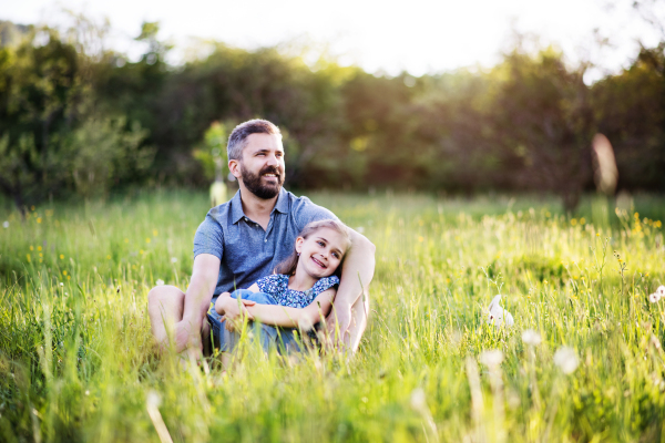 Mature father with a small daughter sitting on the grass in sunny spring nature. Copy space.