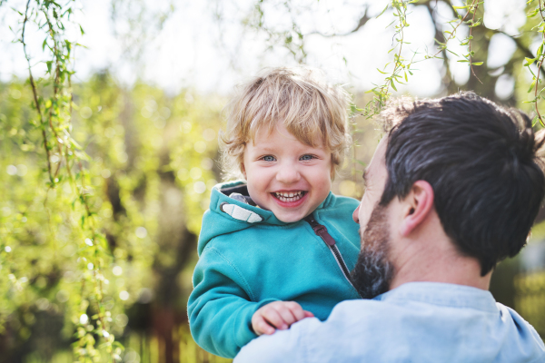 An unrecognizable father with his toddler son outside in green sunny spring nature, having fun.