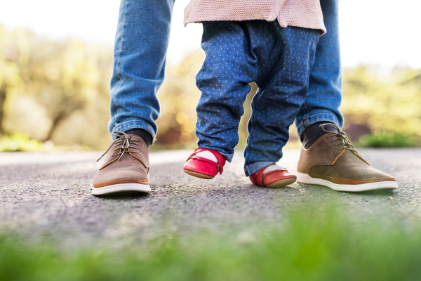 First steps of a toddler girl outside in spring nature. Unrecognizable father holding his daughter when walking.