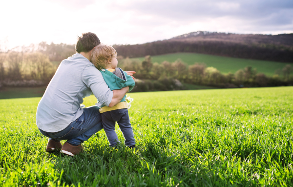 A father with his toddler son outside in green sunny spring nature. Rear view. Copy space.