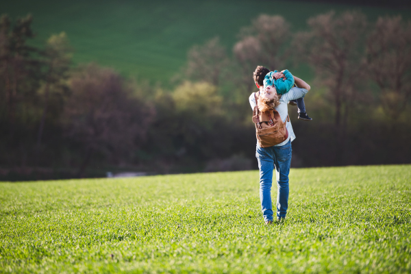 A father with his toddler son on a walk outside in green sunny spring nature, having fun. Rear view.