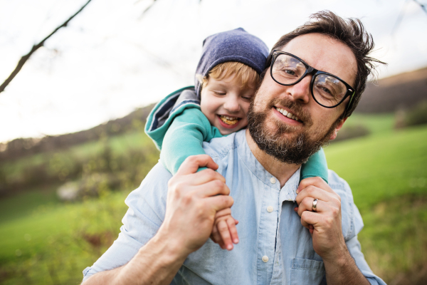 A father holding his toddler son on back outside in green sunny spring nature.