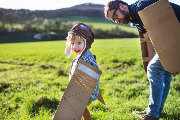 Happy toddler boy with hat, goggles and wings playing outside with father in spring nature. Pilot and flying concept.