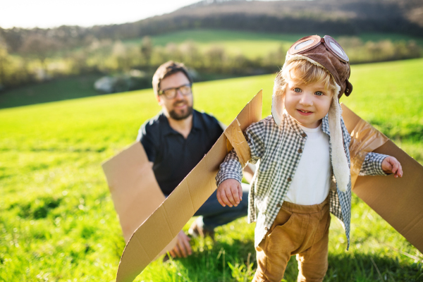 Happy toddler boy with hat, goggles and wings playing outside with father in spring nature. Pilot and flying concept.