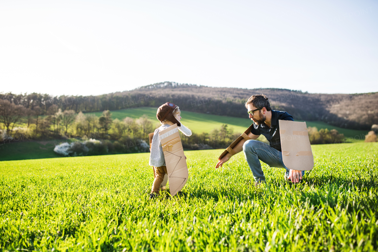 Happy toddler boy with hat, goggles and wings playing outside with father in spring nature. Pilot and flying concept.