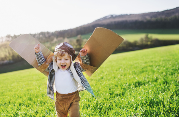 Happy toddler boy with hat, goggles and wings playing outside in spring nature. Pilot and flying concept.