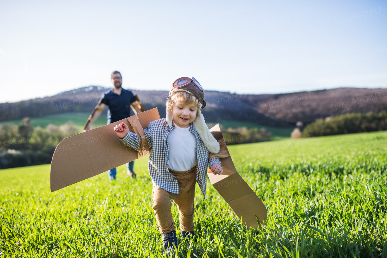 Happy toddler boy with hat, goggles and wings playing outside with father in spring nature. Pilot and flying concept.