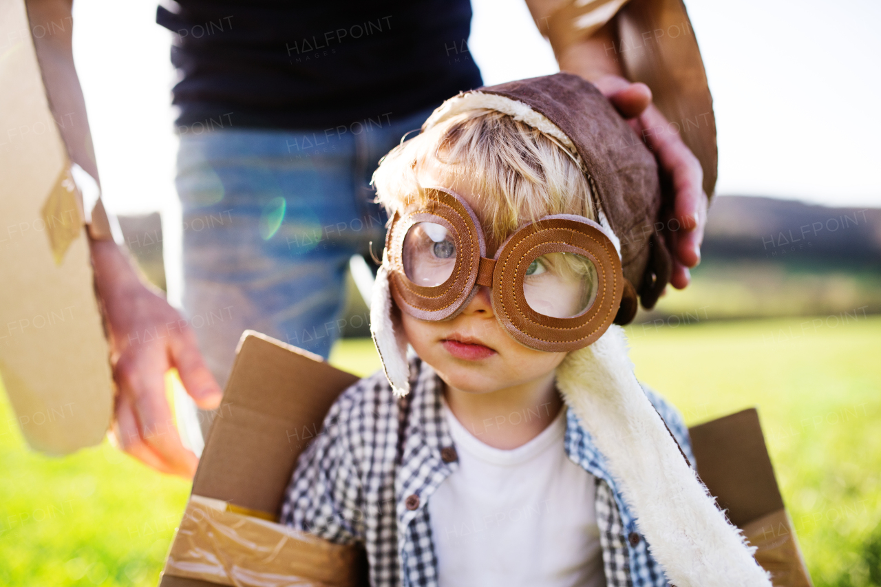 Happy toddler boy with hat, goggles and wings playing outside with unrecognizable father in spring nature. Pilot and flying concept.