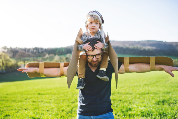 Happy toddler boy with hat, goggles and wings playing outside with his father in spring nature. Pilot and flying concept.