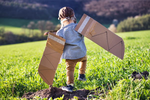 Happy toddler boy with hat, goggles and wings playing outside in spring nature. Pilot and flying concept. Rear view.