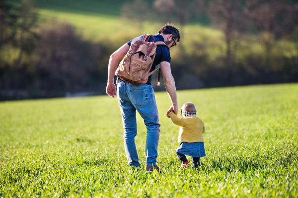 A father with his toddler daughter on a walk outside in green sunny spring nature. Rear view.