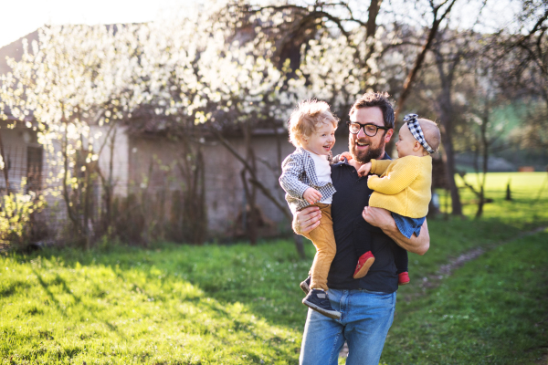 A father holding his toddler children outside in green sunny spring nature. A handsome man with little girl and boy having fun.