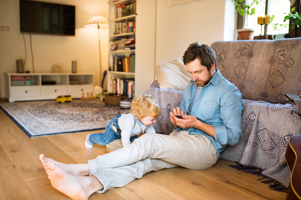 Young father at home, holding smart phone, texting, his cute little son playing around him