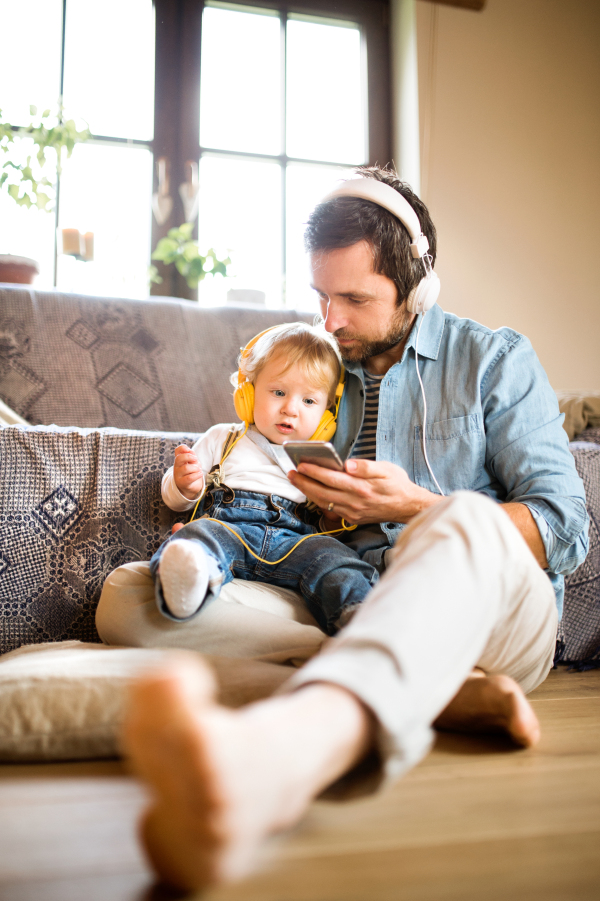 Young father and his little son with smart phone and earphones, listening music at home, sitting on wooden floor.