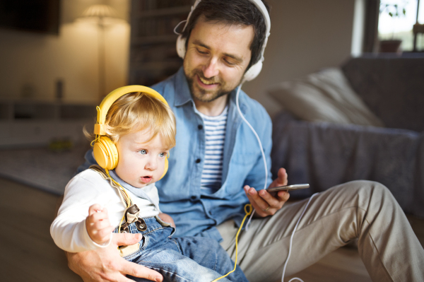 Young father and his little son with smart phone and earphones, listening music at home, sitting on wooden floor.