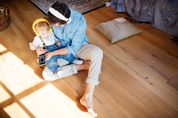 Young father and his little son with smart phone and earphones, listening music at home, sitting on wooden floor.