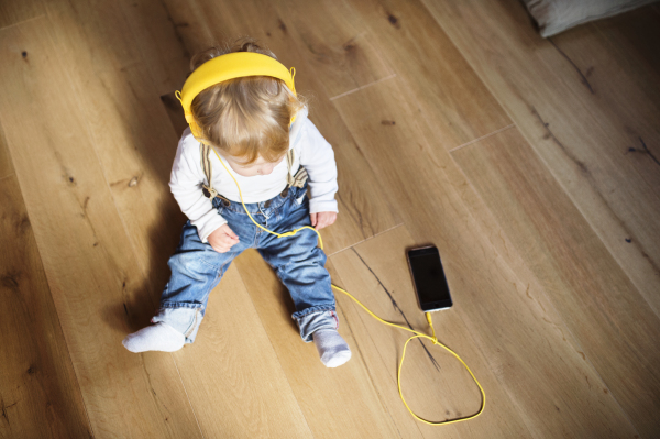 Cute little boy at home sitting on wooden floor wearing headphones, listening music from smart phone. High angle view.