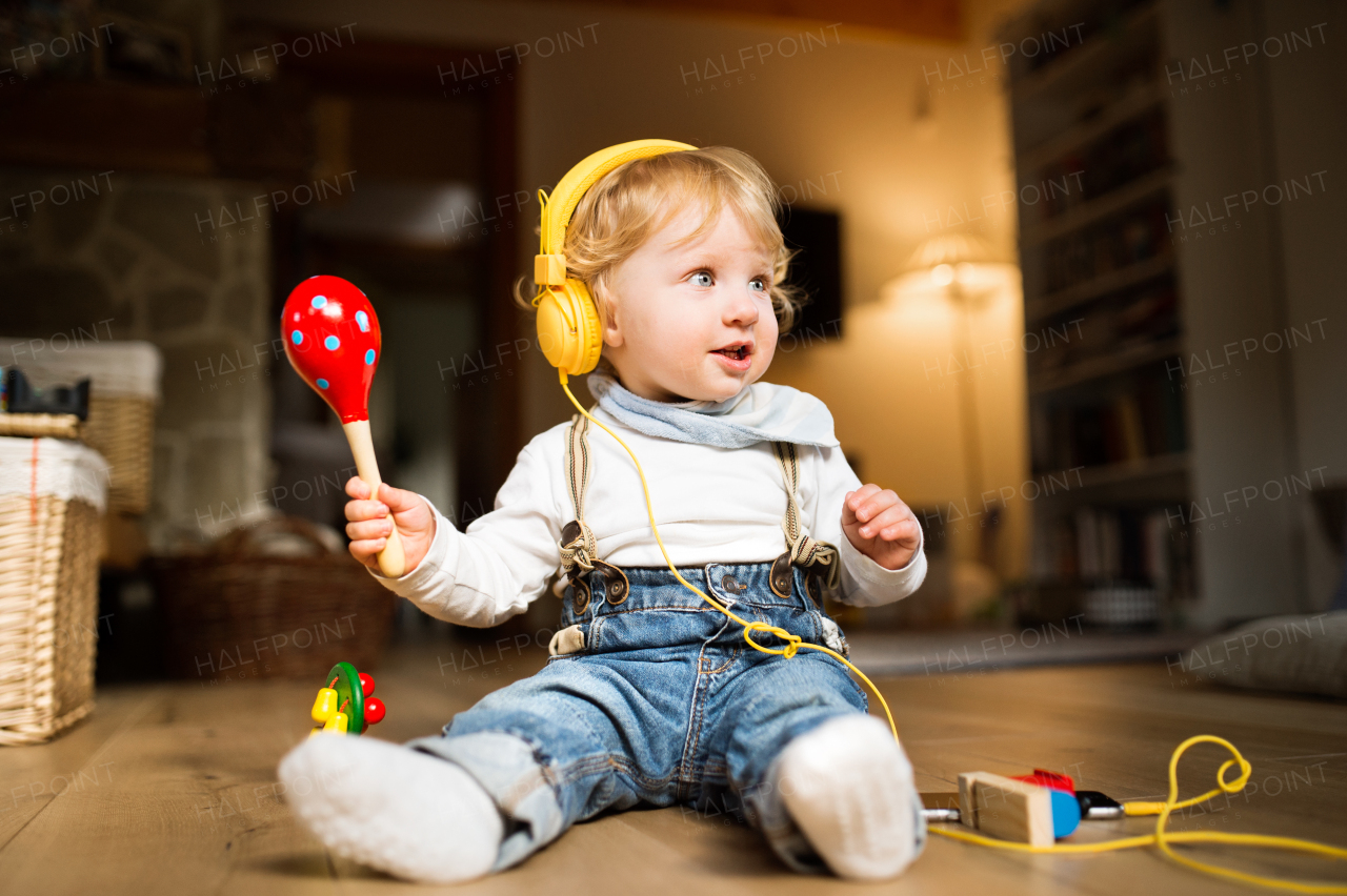 Cute little boy at home sitting on wooden floor wearing earphones, listening music from smart phone and plying musical instrument.