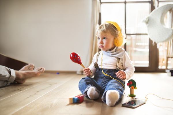 Cute little boy at home sitting on wooden floor wearing headphones, listening music from smart phone and playing musical instrument.