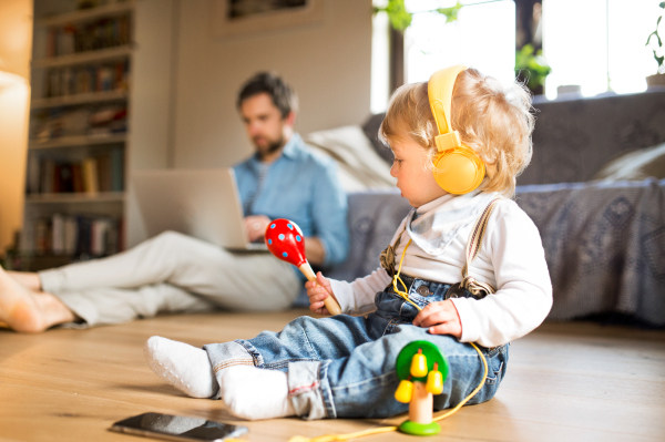 Young father at home sitting on wooden floor, working on laptop, his cute little son with smart phone and earphones, listening music, playing with musical instruments.