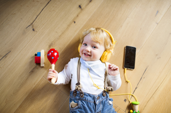 Cute little boy at home lying on wooden floor wearing headphones, listening music from smart phone and playing musical instrument.