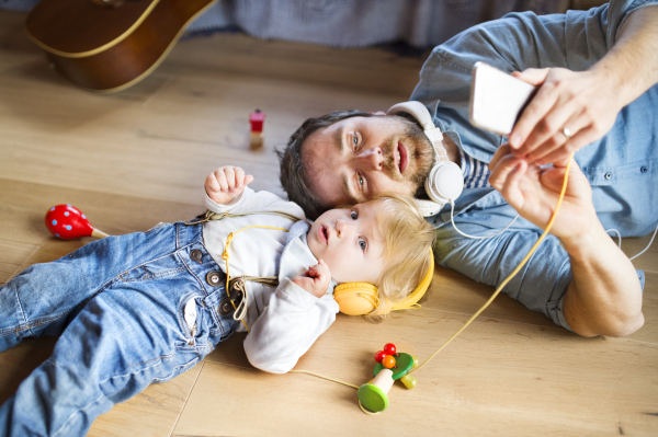 Young father and his little son with smart phone and earphones, listening music at home, lying on wooden floor, musical toys surrounding them.