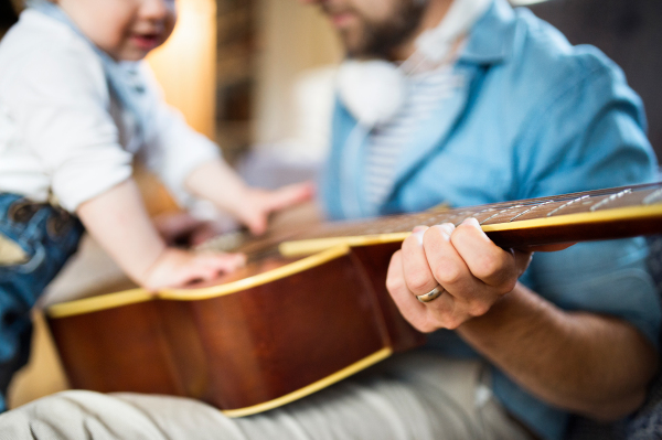 Unrecognizable young father with earphones at home and his little son trying to play guitar.