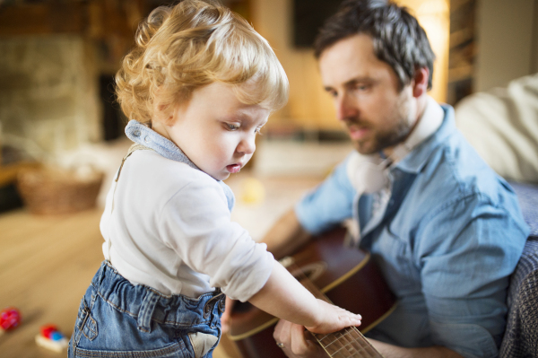 Young father at home sitting on wooden floor, his cute little son playing guitar that he is holding.