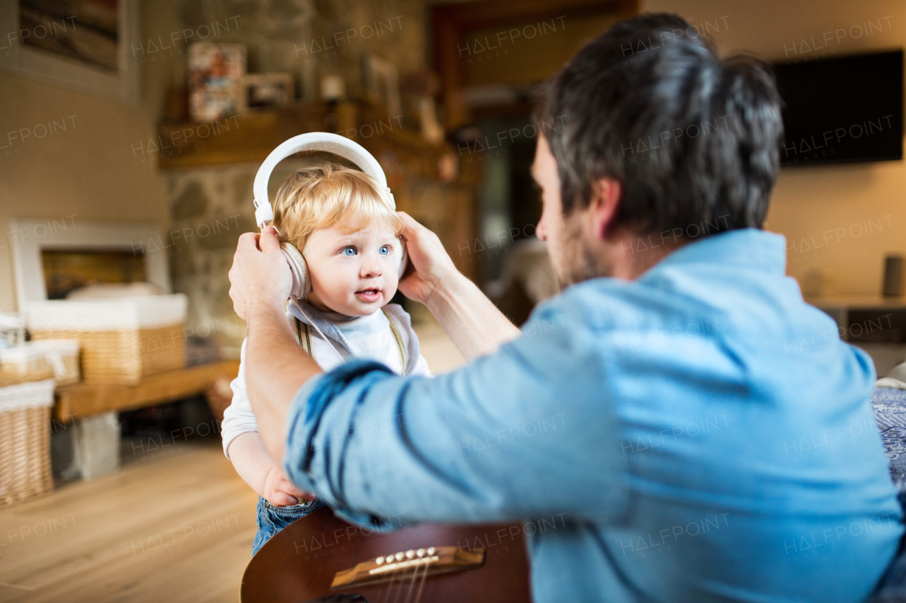 Unrecognizable young father at home with guitar, his little son wearing earphones and listening music.