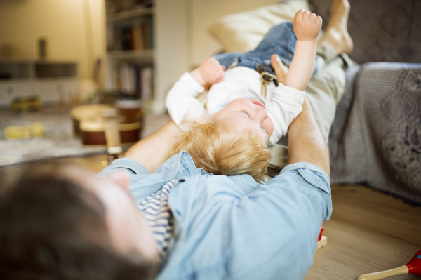 Young father at home lying on wooden floor playing with his little son, cuddling him.