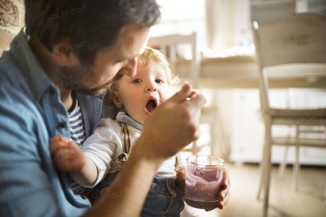 Young father at home sitting on the floor feeding his cute little son with yoghurt.