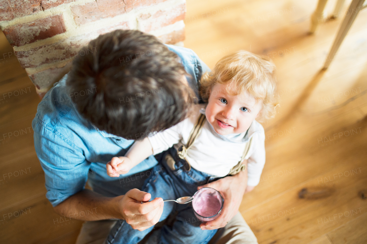 Young father at home sitting on the floor feeding his cute little son with yoghurt.