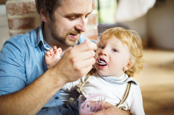 Young father at home sitting on the floor feeding his cute little son with yoghurt.