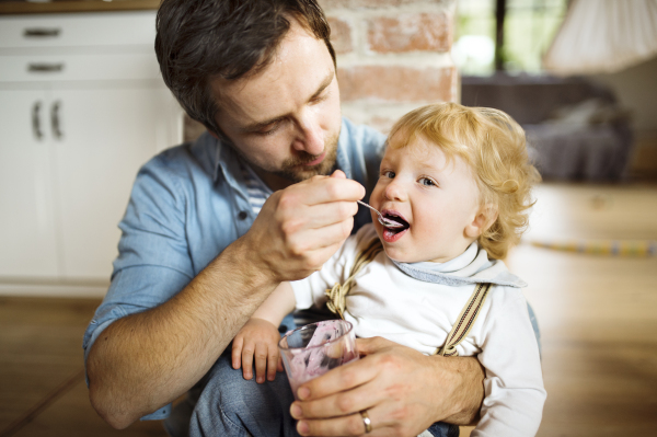 Young father at home sitting on the floor feeding his cute little son with yoghurt.