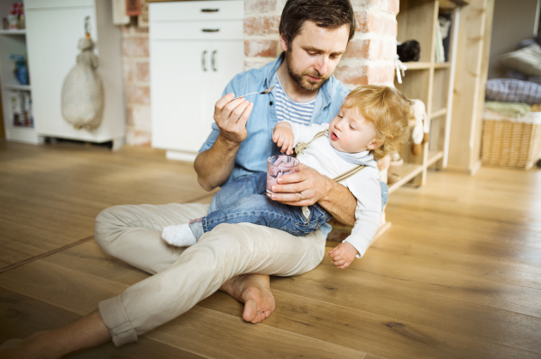 Young father at home sitting on the floor feeding his cute little son with yoghurt.