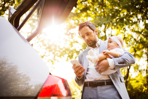 Young father holding his little baby girl in the arms going into the car. Man with smartphone texting.