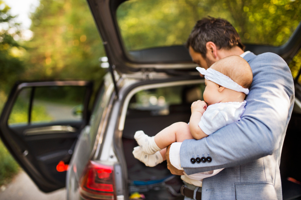 Young father holding his little baby girl standing by the car. Doors opened.