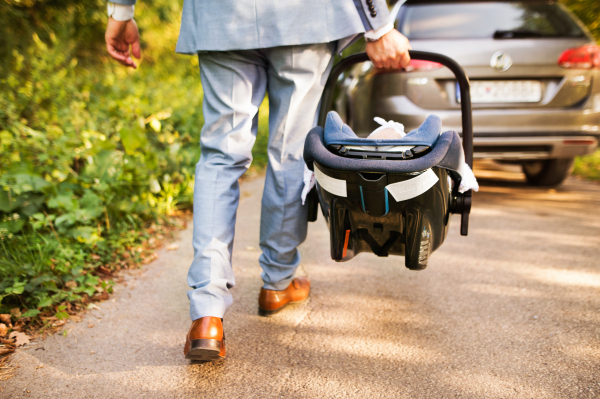 Unrecognizable young father carrying his little baby in a car seat, going into the car.