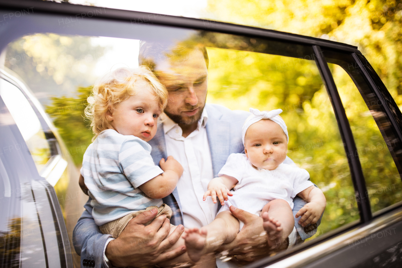 Young father with baby and toddler by the car. Man carrying baby girl and toddler boy. Shot through glass.