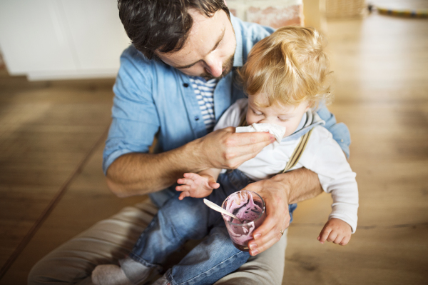 Young father at home sitting on the floor feeding his cute little son with yoghurt, wiping his face with napkin.