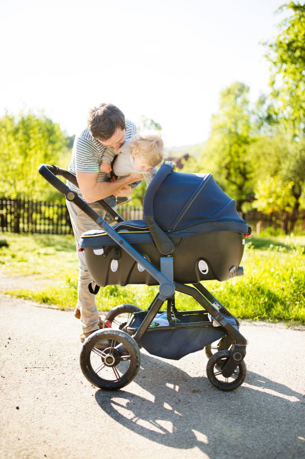 Young father outdoors in sunny summer park holding his cute little son in the arms looking at baby girl lying in stroller.