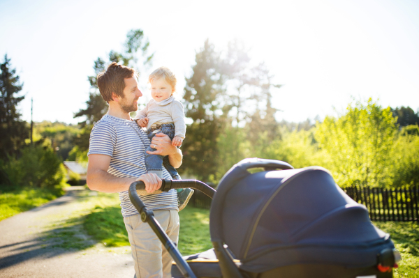 Young father outdoors on a walk in sunny summer park holding his cute little son in the arms, baby girl lying in stroller.