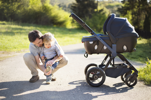 Young father outdoors in sunny summer park with cute little son and baby daughter in stroller.