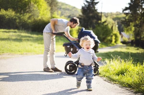 Young father outdoors in sunny summer park with cute little son and baby daughter in stroller.