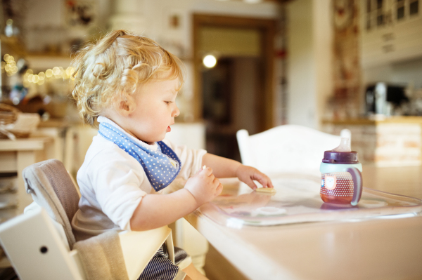 Cute little blonde boy at home sitting at the table, eating and drinking.