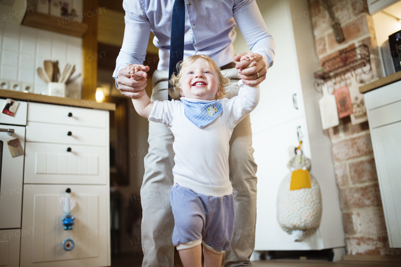 Unrecognizable father coming home from the office, holding hands of his little son taking first steps.